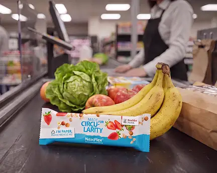 Snackriegel in recycelbarer Papierverpackung mit Aufdruck "Made for Circularity" an Supermarktkasse neben Bananen, Salat und &Auml;pfeln.