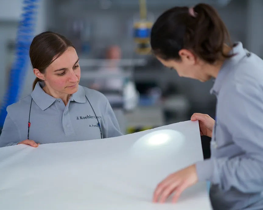 Zwei Mitarbeiterinnen im Koehler-Werk pr&uuml;fen ein gro&szlig;es wei&szlig;es Papierbahn-Muster mit hellem Pr&uuml;flicht im Laborbereich.
