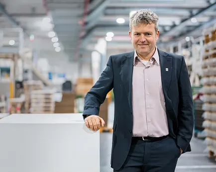 Jens Kriete im dunklen Anzug lehnt an gestapelten Papierb&ouml;gen in moderner Produktionshalle des Unternehmens.