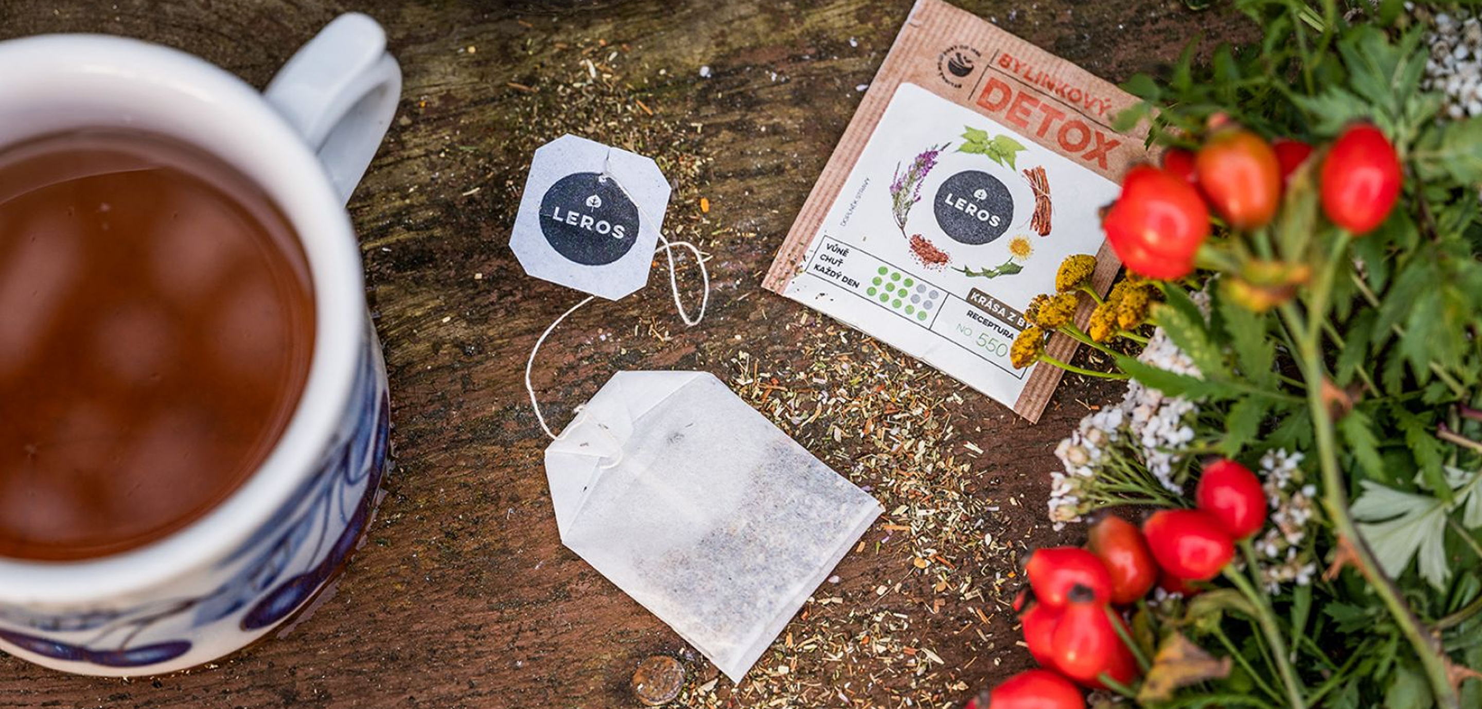 Tasse Kr&auml;utertee mit LEROS-Teebeutel, Detox-Packung und frischen roten Beeren sowie Kr&auml;utern auf rustikalem Holztisch