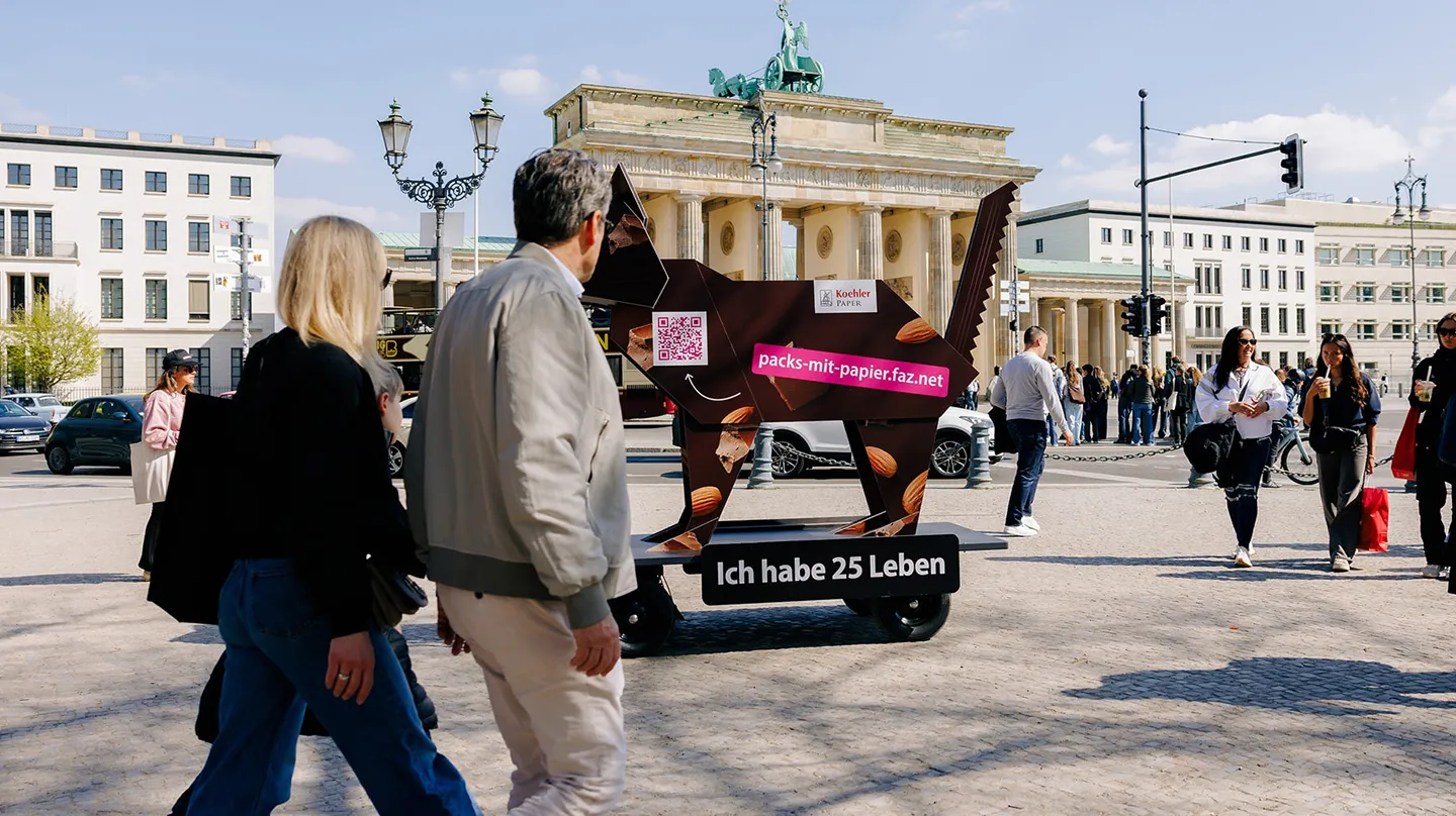 Passantinnen und Passanten vor  Origami-Katzen-Figur mit QR-Code und URL packs-mit-papier.faz.net, dahinter Brandenburger Tor