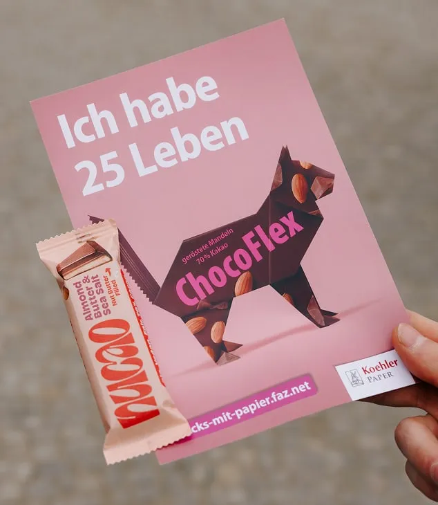 Hand h&auml;lt pinken Flyer zur Kampagne "Pack's mit Papier" mit Slogan "Ich habe 25 Leben", davor Nucao-Riegel Almond Butter & Sea Salt