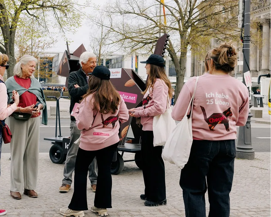 Promoteam in rosa Kampagnen-Pullovern mit "Ich habe 25 Leben" Slogans spricht mit Passantinnen vor Origami-Katze auf Anh&auml;nger in Berlin