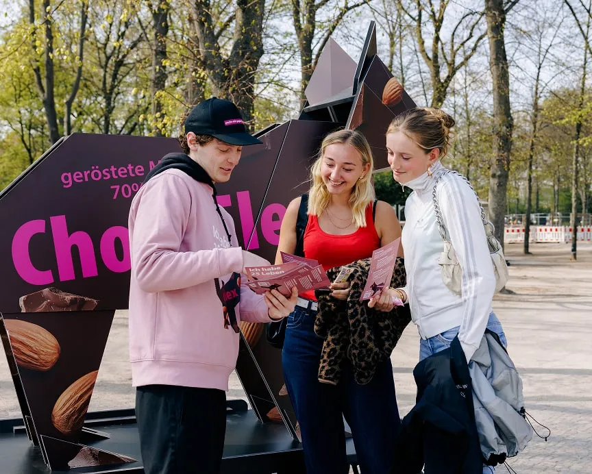 Junger Promoter im rosa Hoodie gibt zwei Frauen Flyer zur Kampagne "Pack's mit Papier" mit Slogan "Ich habe 25 Leben" vor gro&szlig;er Origami-Katze